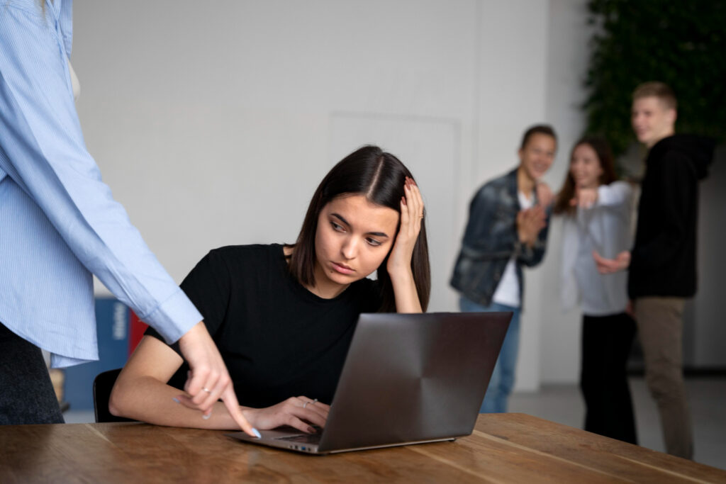 Una mujer joven, sentada frente a un ordenador portátil, muestra signos de preocupación mientras una persona le señala algo de manera autoritaria. Al fondo, tres compañeros se ríen y apuntan hacia ella, representando una situación de acoso laboral o mobbing en el entorno de trabajo.